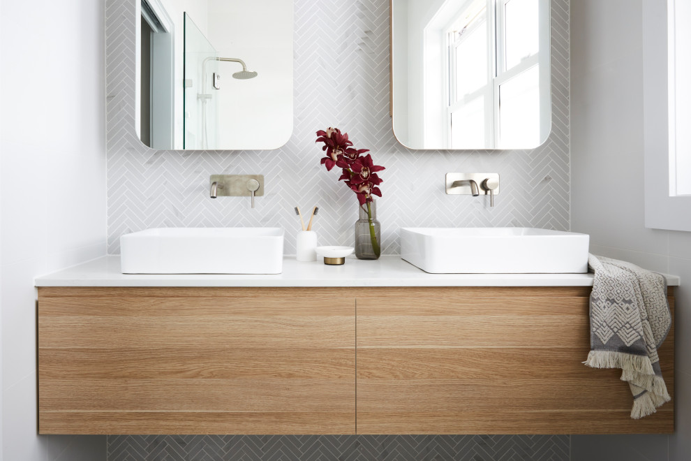 Nordisk inredning av ett vit vitt badrum, med skåp i ljust trä, ett fristående badkar, en toalettstol med hel cisternkåpa, vit kakel, keramikplattor, vita väggar, klinkergolv i keramik, marmorbänkskiva och grått golv