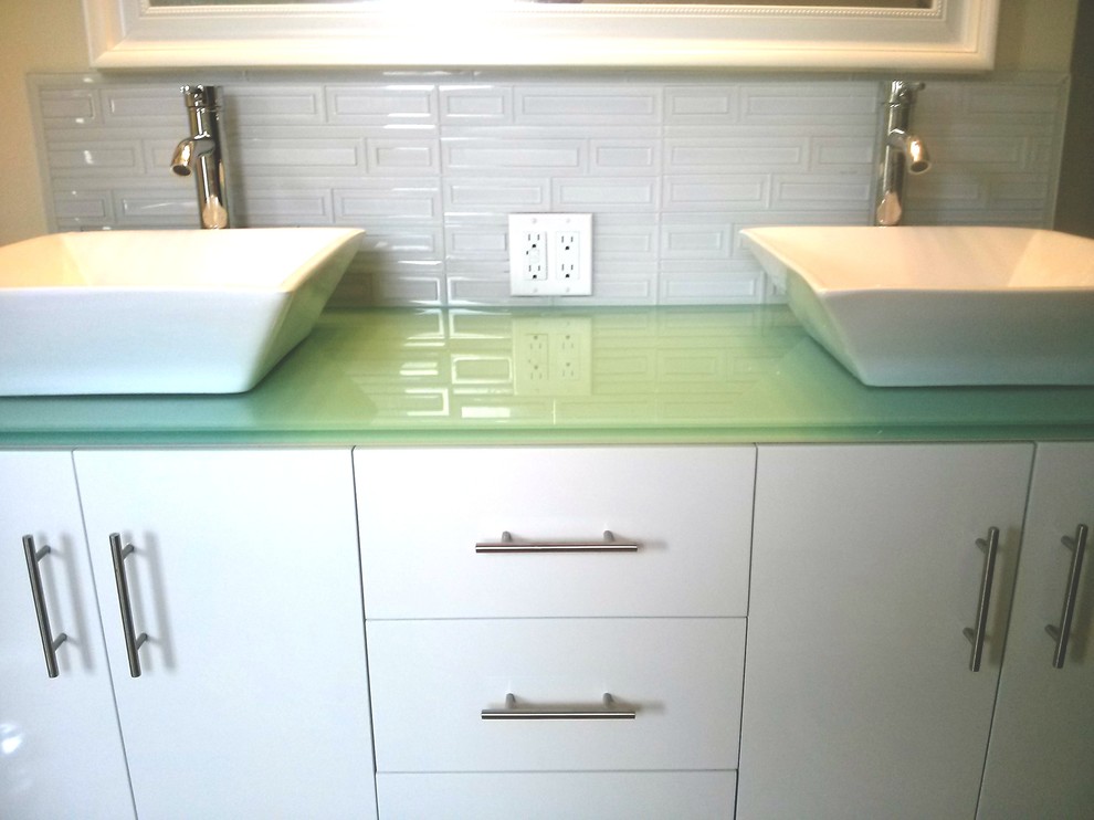 Cette photo montre une salle de bain bord de mer de taille moyenne avec un placard à porte plane, des portes de placard blanches, un carrelage vert, un mur blanc, une vasque et un plan de toilette en verre.