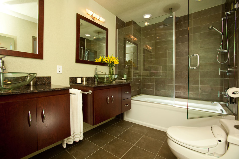 Inredning av ett modernt badrum, med ett fristående handfat, släta luckor, skåp i mörkt trä, ett badkar i en alkov, en dusch/badkar-kombination, en toalettstol med hel cisternkåpa, brun kakel, beige väggar och marmorbänkskiva