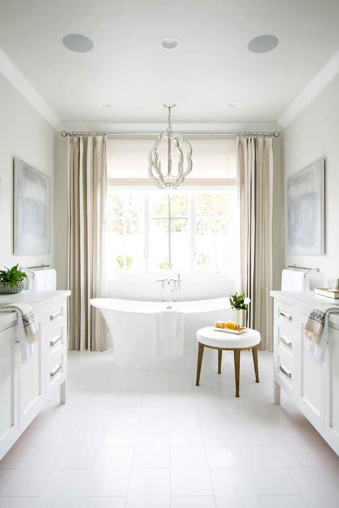 Modelo de cuarto de baño costero con armarios con paneles empotrados, puertas de armario blancas, bañera exenta, paredes beige, suelo blanco y encimeras blancas