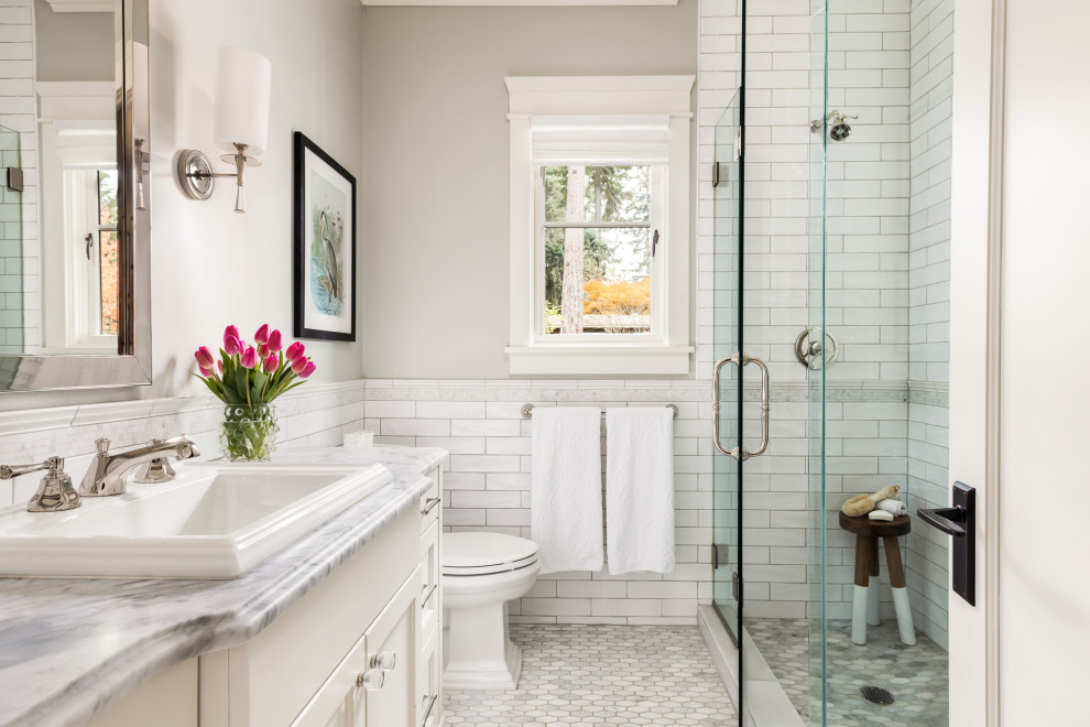 Foto på ett vintage grå badrum med dusch, med luckor med infälld panel, vita skåp, en dusch i en alkov, en toalettstol med separat cisternkåpa, vit kakel, tunnelbanekakel, vita väggar, ett nedsänkt handfat, marmorbänkskiva, grått golv och dusch med gångjärnsdörr