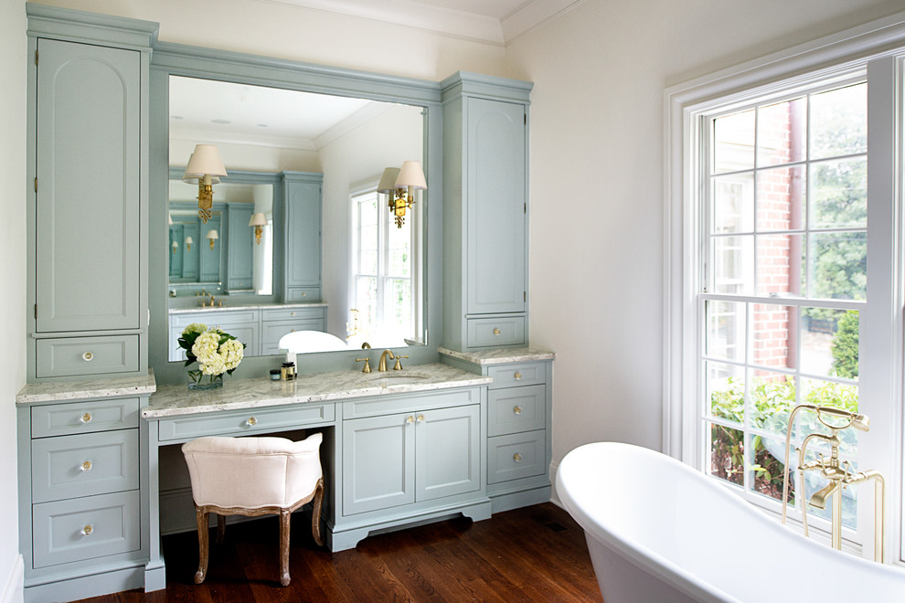 Foto på ett vintage en-suite badrum, med blå skåp, granitbänkskiva, mörkt trägolv, ett undermonterad handfat, ett badkar med tassar, vita väggar och luckor med infälld panel