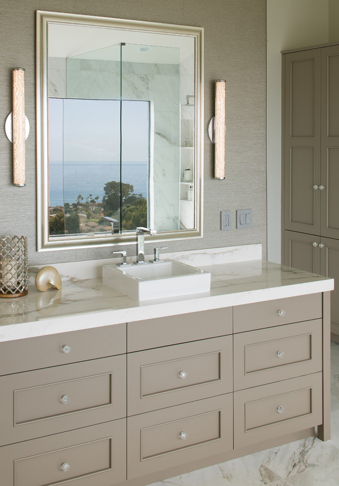 Cette photo montre une salle de bain principale bord de mer avec un mur gris, un sol en marbre, une vasque, un plan de toilette en marbre, un plan de toilette blanc, des portes de placard marrons, un sol blanc et un placard avec porte à panneau encastré.