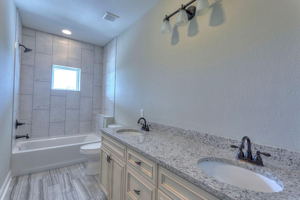 Klassisk inredning av ett mellanstort grå grått en-suite badrum, med luckor med upphöjd panel, vita skåp, ett badkar i en alkov, en dusch/badkar-kombination, en toalettstol med separat cisternkåpa, grå kakel, porslinskakel, grå väggar, klinkergolv i porslin, ett undermonterad handfat, granitbänkskiva och brunt golv