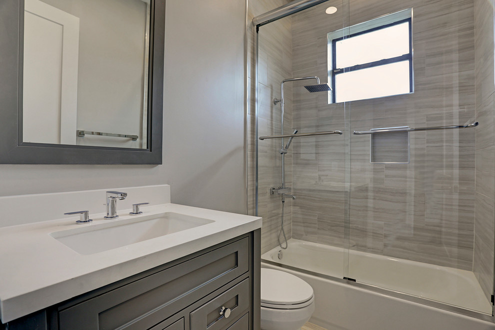 This is an example of a contemporary family bathroom in Houston with shaker cabinets, grey cabinets, a shower/bath combination, a one-piece toilet, beige tiles, ceramic tiles, grey walls, porcelain flooring, a submerged sink, engineered stone worktops, beige floors, a sliding door and white worktops.