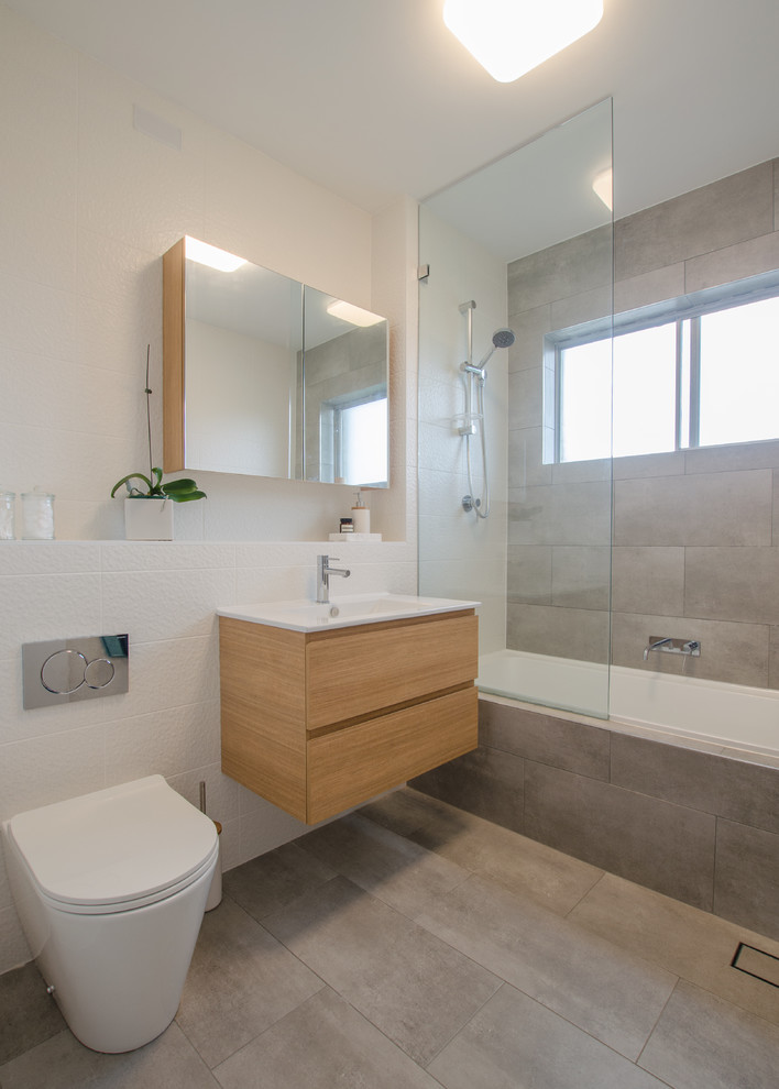 Photo of a small contemporary bathroom in Sydney with flat-panel cabinets, light wood cabinets, a built-in bath, a shower/bath combination, grey tiles, ceramic tiles, engineered stone worktops, a two-piece toilet, white walls, cement flooring and a built-in sink.