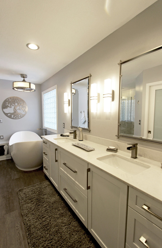 Пример оригинального дизайна: главная ванная комната среднего размера в стиле неоклассика (современная классика) с плоскими фасадами, белыми фасадами, отдельно стоящей ванной, угловым душем, керамогранитной плиткой, серыми стенами, полом из ламината, врезной раковиной, столешницей из искусственного кварца, коричневым полом и душем с распашными дверями