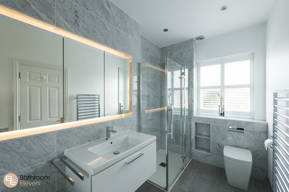 This is an example of a medium sized contemporary bathroom with white cabinets, a built-in bath, grey tiles, white walls, a walk-in shower, a one-piece toilet, porcelain tiles, light hardwood flooring and a wall-mounted sink.