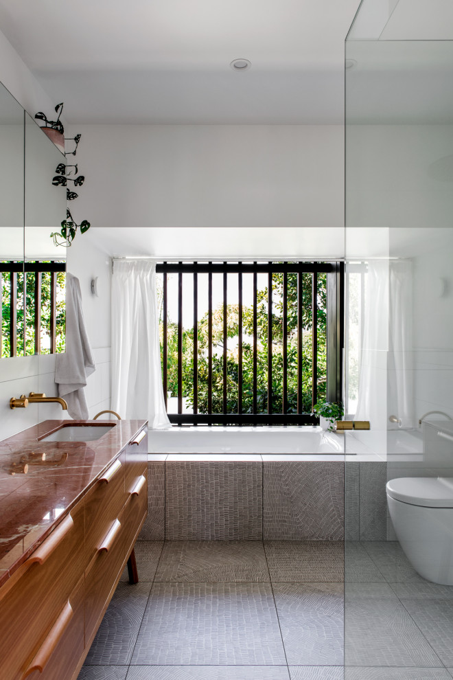 На фото: главная ванная комната среднего размера в стиле фьюжн с фасадами островного типа, светлыми деревянными фасадами, накладной ванной, угловым душем, унитазом-моноблоком, белой плиткой, керамической плиткой, белыми стенами, полом из керамогранита, врезной раковиной, столешницей из гранита, серым полом, душем с распашными дверями, розовой столешницей, тумбой под одну раковину и напольной тумбой с