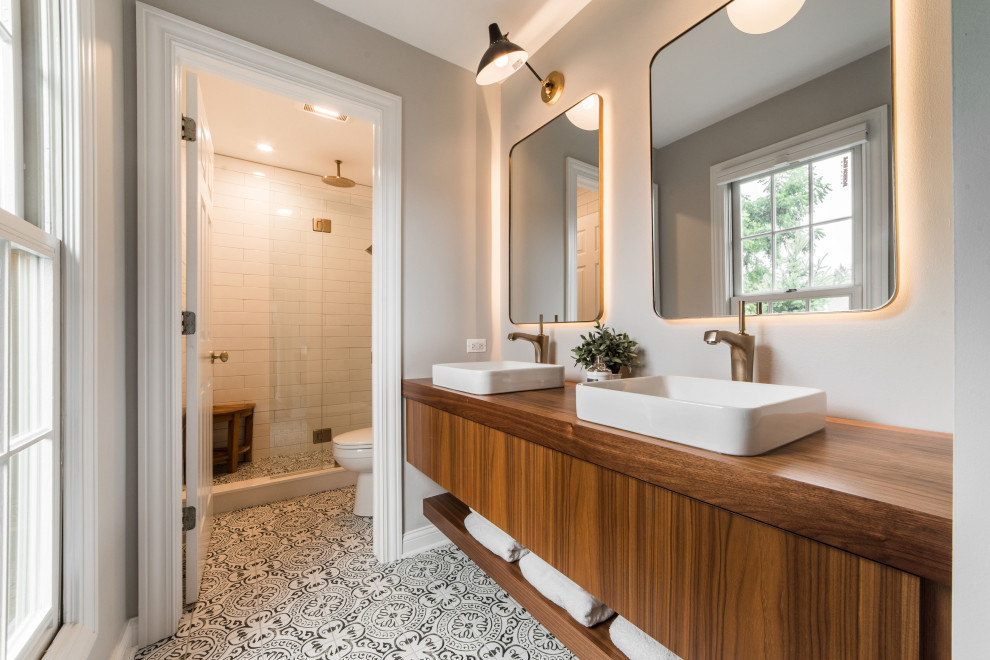 Стильный дизайн: ванная комната в стиле неоклассика (современная классика) с плоскими фасадами, коричневыми фасадами, душем в нише, белой плиткой, серыми стенами, душевой кабиной, настольной раковиной, столешницей из дерева, разноцветным полом, душем с раздвижными дверями, коричневой столешницей, сиденьем для душа, тумбой под две раковины и подвесной тумбой - последний тренд