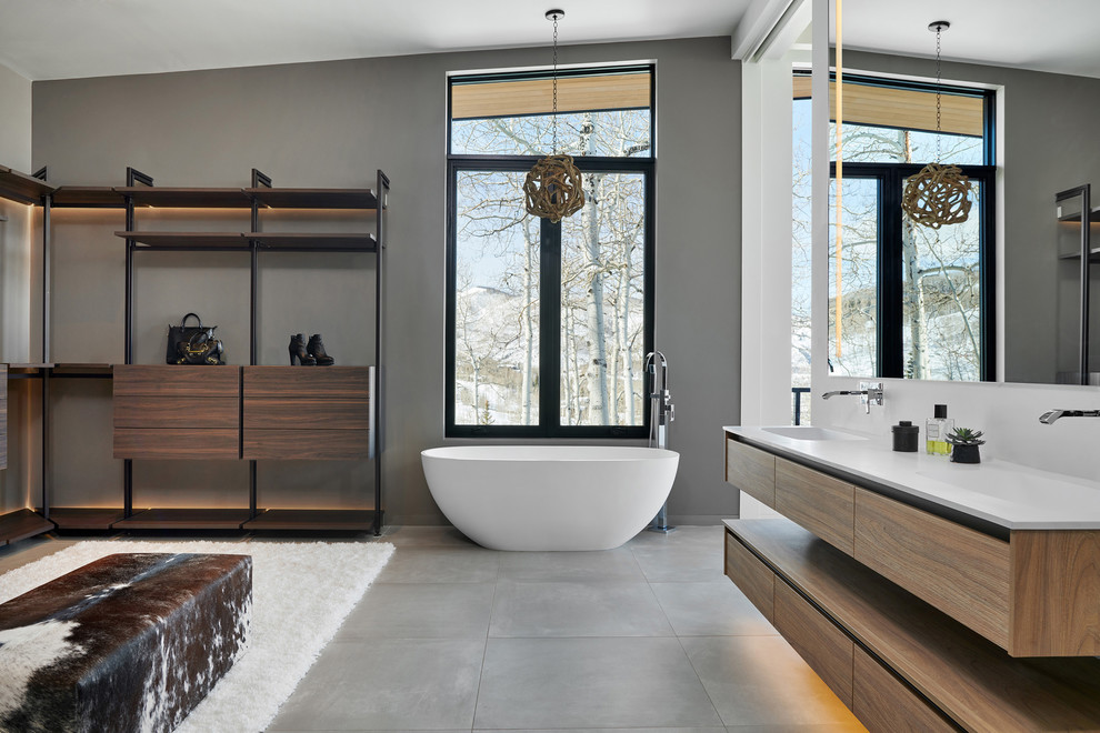 This is an example of a large contemporary ensuite bathroom in Denver with a freestanding bath, porcelain flooring, grey floors, flat-panel cabinets, medium wood cabinets, grey walls, white worktops, a one-piece toilet, grey tiles, cement tiles, an integrated sink and solid surface worktops.