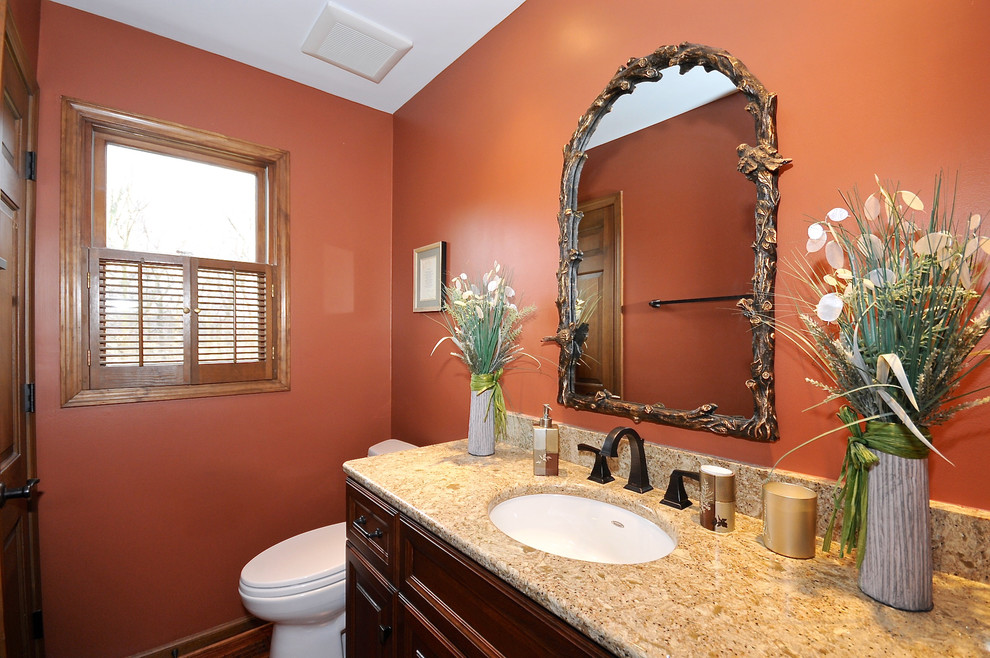 Amerikansk inredning av ett litet badrum, med ett undermonterad handfat, luckor med upphöjd panel, skåp i mellenmörkt trä, granitbänkskiva, en toalettstol med separat cisternkåpa och orange väggar