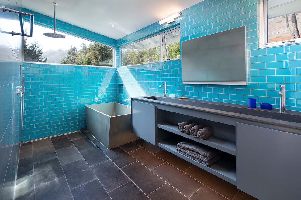 Foto på ett mellanstort funkis grå en-suite badrum, med släta luckor, grå skåp, ett fristående badkar, blå kakel, glaskakel, skiffergolv, ett avlångt handfat, bänkskiva i akrylsten, svart golv, med dusch som är öppen och våtrum
