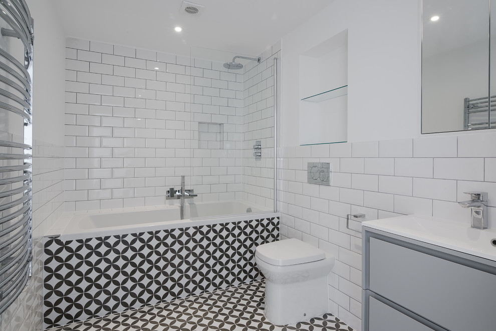 This is an example of a small modern family bathroom in London with flat-panel cabinets, grey cabinets, a built-in bath, a shower/bath combination, a one-piece toilet, white tiles, metro tiles, white walls, porcelain flooring, a built-in sink, black floors and a hinged door.