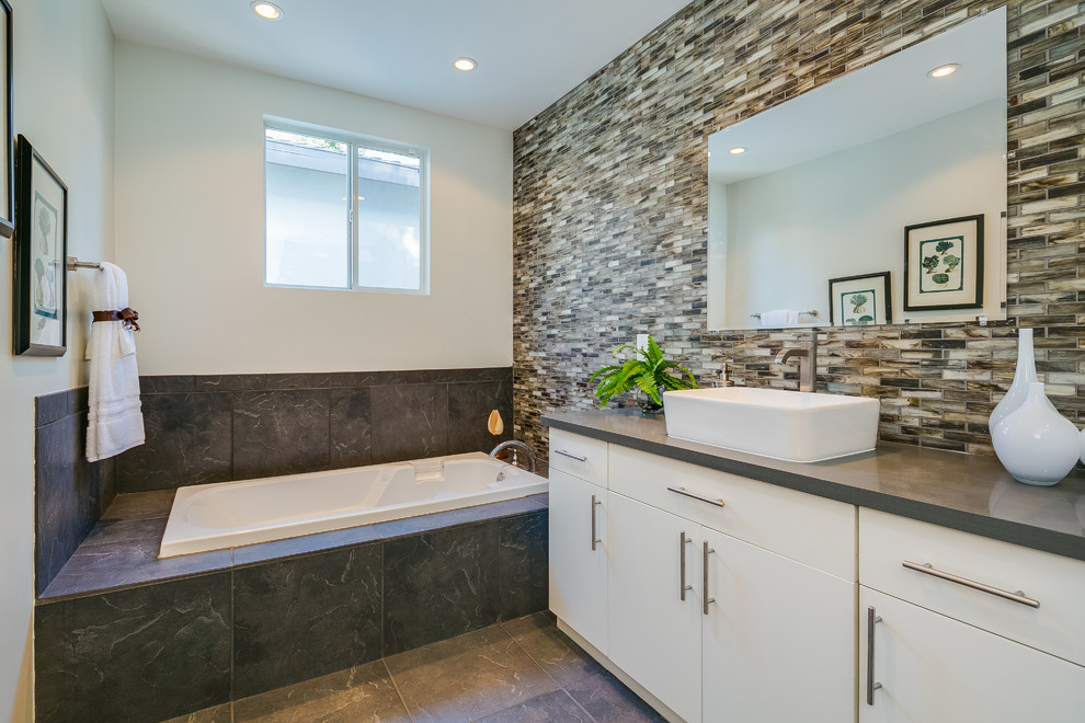Cette photo montre une salle de bain principale tendance avec une vasque, un placard à porte plane, des portes de placard blanches, un plan de toilette en quartz modifié, une baignoire posée, un carrelage gris et un mur gris.