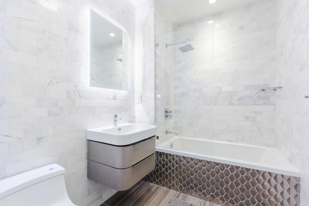 Medium sized scandi bathroom in New York with freestanding cabinets, grey cabinets, a built-in bath, a shower/bath combination, a one-piece toilet, white tiles, marble tiles, porcelain flooring, a wall-mounted sink and an open shower.