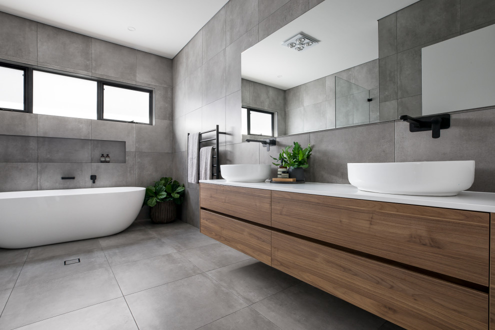 Источник вдохновения для домашнего уюта: ванная комната в современном стиле с плоскими фасадами, фасадами цвета дерева среднего тона, отдельно стоящей ванной, серой плиткой, настольной раковиной, серым полом и белой столешницей