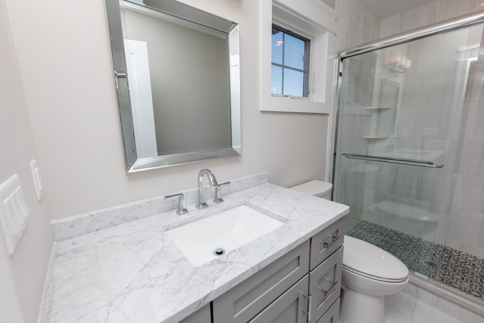 Photo of a coastal bathroom in New York with shaker cabinets, grey cabinets, an alcove shower, a one-piece toilet, grey tiles, ceramic tiles, grey walls, ceramic flooring, a submerged sink, granite worktops, grey floors, a sliding door, white worktops, a single sink and a built in vanity unit.