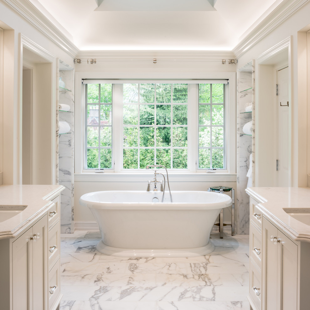 Cette photo montre une salle de bain principale chic avec des portes de placard blanches, une baignoire indépendante, un carrelage blanc, un carrelage de pierre, un sol en marbre, un lavabo encastré, un plan de toilette en quartz, un placard avec porte à panneau encastré et un mur blanc.