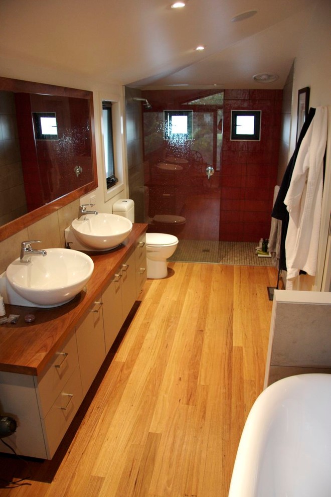 Modern inredning av ett stort badrum, med ett väggmonterat handfat, släta luckor, träbänkskiva, en hörndusch, en toalettstol med hel cisternkåpa, keramikplattor, vita väggar och mellanmörkt trägolv