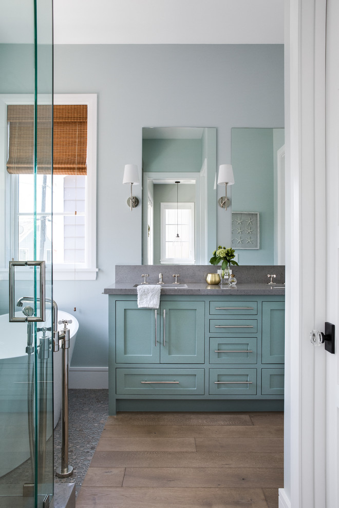 Idée de décoration pour une salle de bain marine avec un placard à porte shaker, des portes de placard bleues, une baignoire indépendante, un mur gris, un sol en bois brun, un lavabo encastré, un sol marron et un plan de toilette gris.