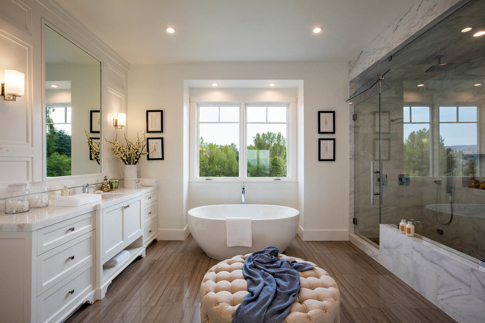 Cette image montre une salle de bain traditionnelle avec un placard à porte shaker, des portes de placard blanches, une baignoire indépendante, un mur blanc, un sol en bois brun, un lavabo encastré, un plan de toilette en marbre et une cabine de douche à porte battante.