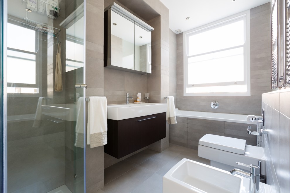 Modernes Badezimmer mit flächenbündigen Schrankfronten, dunklen Holzschränken, Einbaubadewanne, Bidet, grauen Fliesen und Trogwaschbecken in London