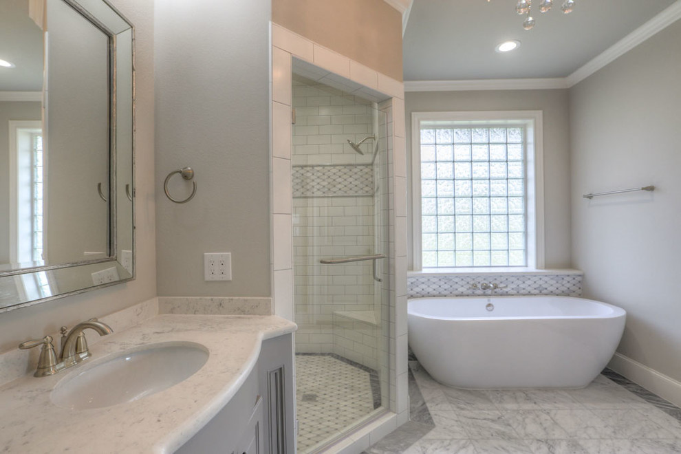 Photo of a large classic ensuite bathroom in Houston with raised-panel cabinets, grey cabinets, a freestanding bath, an alcove shower, a two-piece toilet, grey tiles, mosaic tiles, beige walls, marble flooring, a submerged sink, marble worktops, white floors and a hinged door.