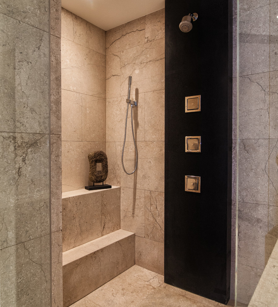 На фото: огромная главная ванная комната в стиле модернизм с плоскими фасадами, душем в нише, черными фасадами, угловой ванной, бежевой плиткой, мраморной плиткой, бежевыми стенами, мраморным полом, врезной раковиной, столешницей из известняка, бежевым полом, душем с распашными дверями и черной столешницей с