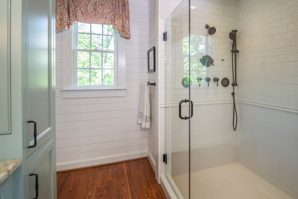 This is an example of a medium sized farmhouse ensuite bathroom in Charlotte with beaded cabinets, an alcove bath, white tiles, white walls, medium hardwood flooring, a built-in sink, quartz worktops, a hinged door and multi-coloured worktops.