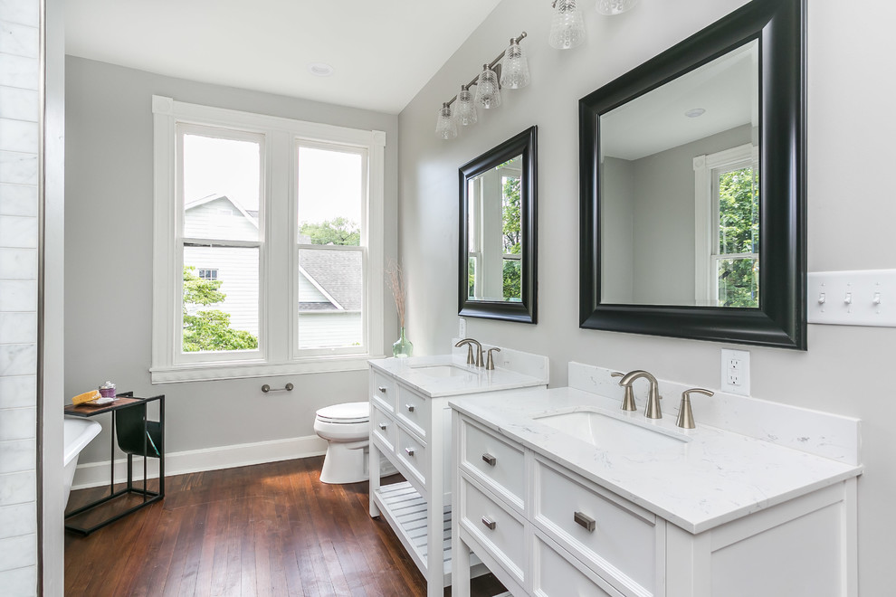 Bild på ett vintage en-suite badrum, med vita skåp, ett fristående badkar, en dusch i en alkov, en toalettstol med separat cisternkåpa, grå kakel, grå väggar, mörkt trägolv, ett konsol handfat, brunt golv och luckor med infälld panel
