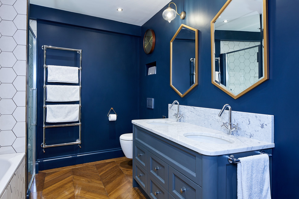 Bild på ett litet funkis badrum för barn, med blå skåp, ett platsbyggt badkar, en öppen dusch, en vägghängd toalettstol, vit kakel, mosaik, blå väggar, ljust trägolv, ett nedsänkt handfat, marmorbänkskiva, brunt golv, med dusch som är öppen och luckor med infälld panel