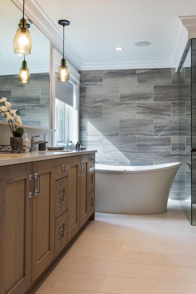 Photo of a large coastal ensuite bathroom in Vancouver with shaker cabinets, brown cabinets, a freestanding bath, an alcove shower, grey tiles, porcelain tiles, grey walls, porcelain flooring, a submerged sink, quartz worktops, beige floors, a hinged door and grey worktops.