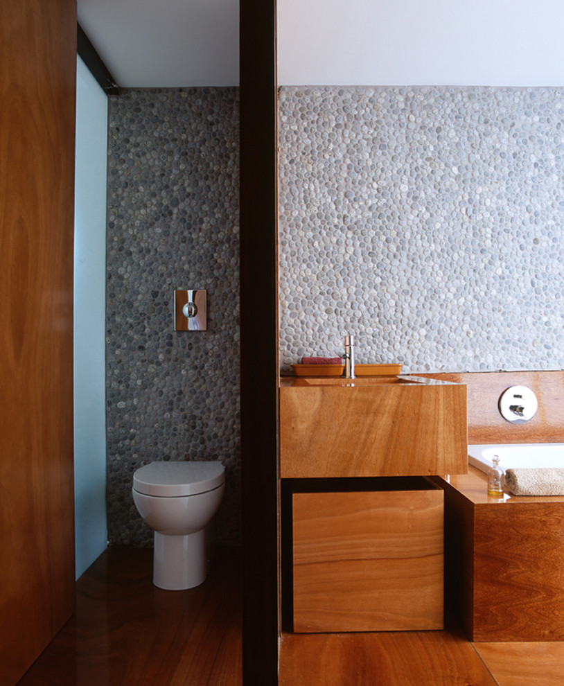Modernes Badezimmer mit flächenbündigen Schrankfronten, hellbraunen Holzschränken, Einbaubadewanne, grauen Fliesen, Kieselfliesen und WC-Raum in Sonstige