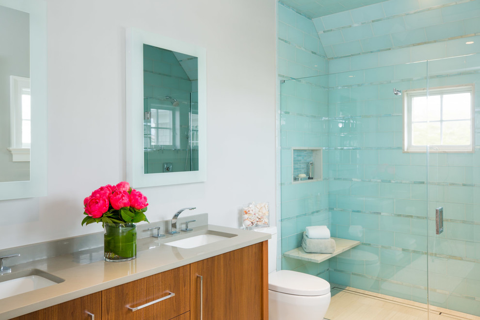 Aménagement d'une salle de bain bord de mer en bois brun avec un placard à porte plane, un carrelage bleu, un mur blanc, un lavabo encastré, une cabine de douche à porte battante, un plan de toilette gris et une fenêtre.