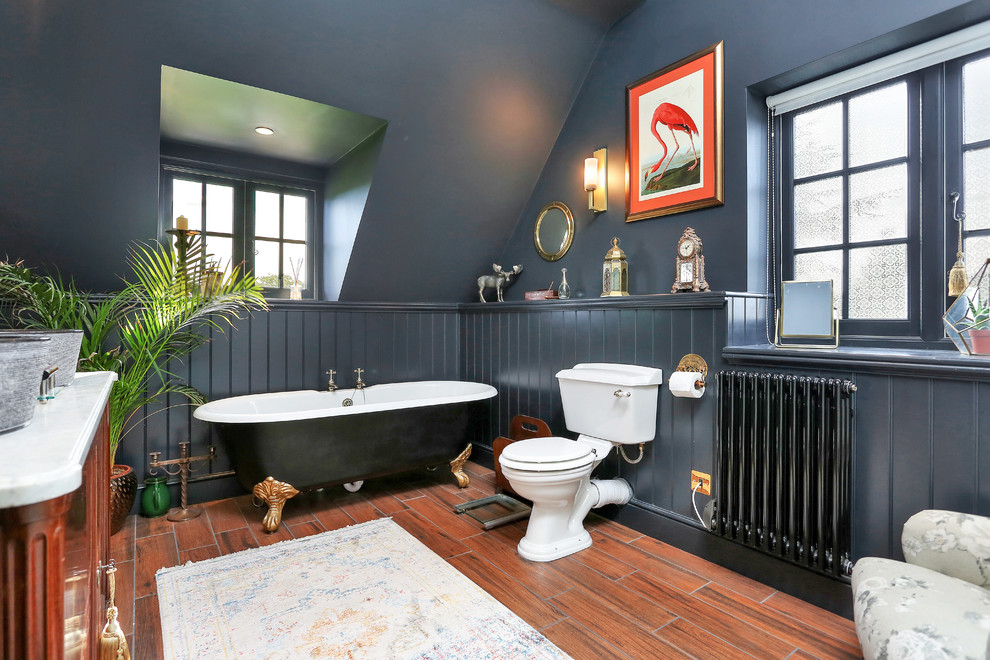 Klassisches Badezimmer mit dunklen Holzschränken, Löwenfuß-Badewanne, Wandtoilette mit Spülkasten, schwarzer Wandfarbe, braunem Holzboden, Aufsatzwaschbecken, braunem Boden und weißer Waschtischplatte in Cambridgeshire