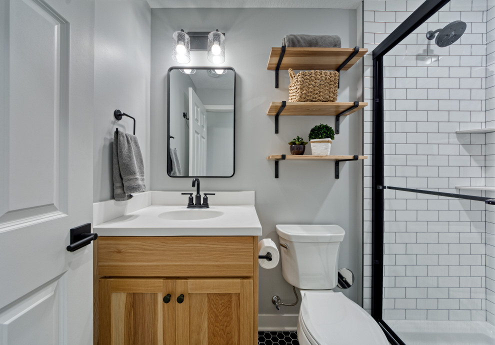 Пример оригинального дизайна: маленькая ванная комната в стиле кантри с плоскими фасадами, светлыми деревянными фасадами, душем в нише, раздельным унитазом, белой плиткой, плиткой кабанчик, серыми стенами, полом из керамической плитки, душевой кабиной, монолитной раковиной, столешницей из искусственного камня, черным полом, душем с раздвижными дверями, белой столешницей, тумбой под одну раковину и встроенной тумбой для на участке и в саду