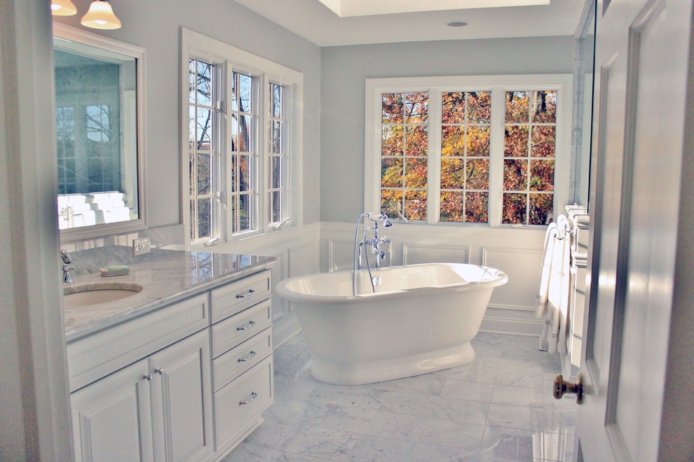 This is an example of a large traditional ensuite half tiled bathroom in Philadelphia with recessed-panel cabinets, white cabinets, a freestanding bath, a corner shower, a two-piece toilet, white tiles, grey walls, marble flooring, a submerged sink and marble worktops.