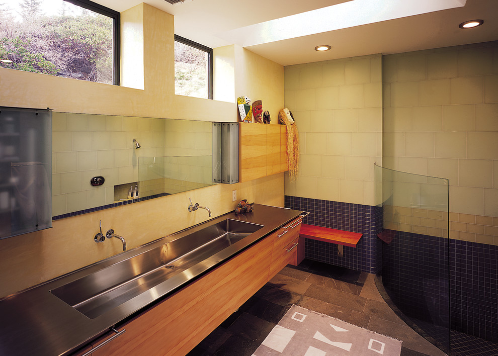 Bild på ett stort funkis badrum, med ett avlångt handfat, släta luckor, skåp i ljust trä, bänkskiva i rostfritt stål, en öppen dusch, grön kakel, glaskakel, gula väggar och skiffergolv