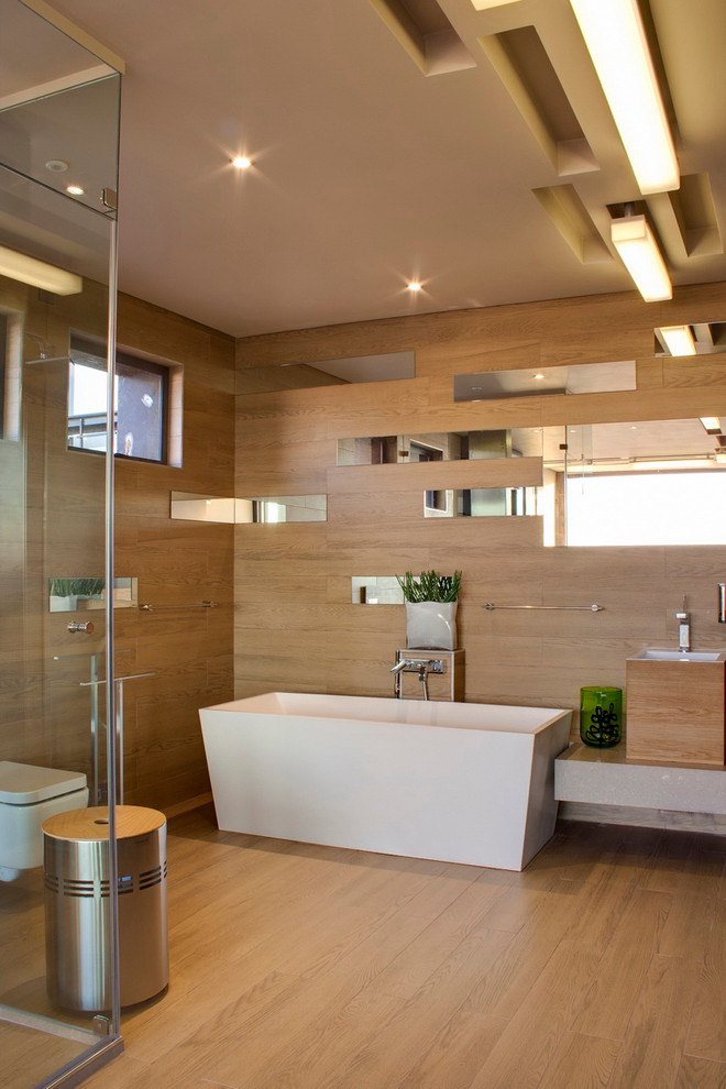 Modernes Badezimmer mit Wandwaschbecken, freistehender Badewanne, Wandtoilette, Spiegelfliesen, beiger Wandfarbe und braunem Holzboden in Sonstige