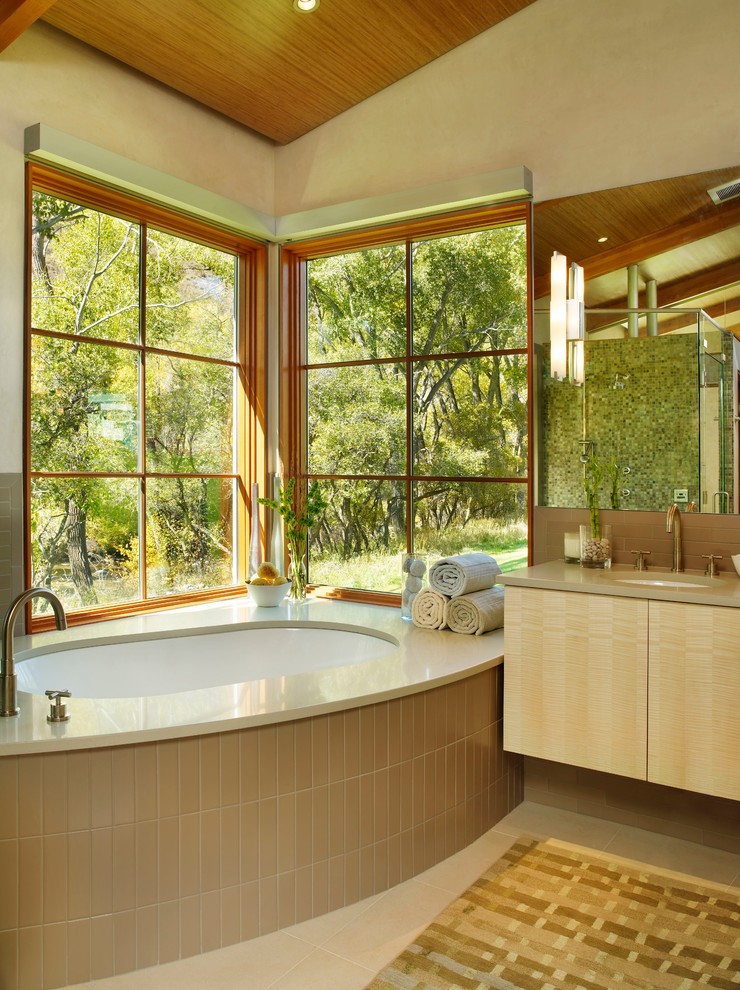Immagine di una stanza da bagno contemporanea con lavabo sottopiano, ante lisce, ante in legno chiaro, vasca ad angolo, piastrelle beige e pareti beige