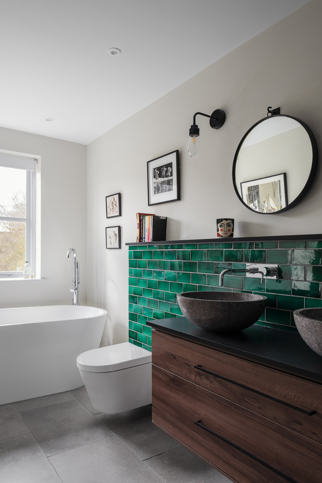 Exemple d'une salle de bain tendance en bois foncé avec un placard à porte plane, une baignoire indépendante, un carrelage vert, un mur gris, une vasque, un sol gris, un plan de toilette noir, meuble double vasque et meuble-lavabo suspendu.