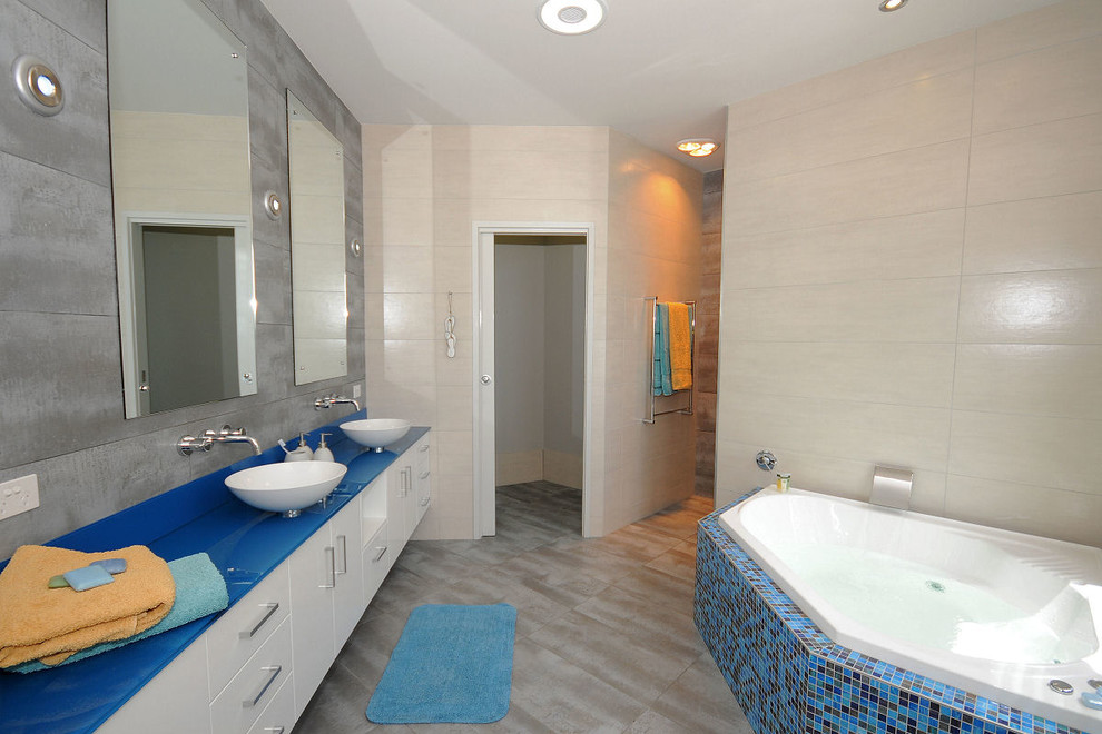 Modern inredning av ett badrum, med ett fristående handfat, släta luckor, vita skåp, ett hörnbadkar, blå kakel, mosaik och beige väggar