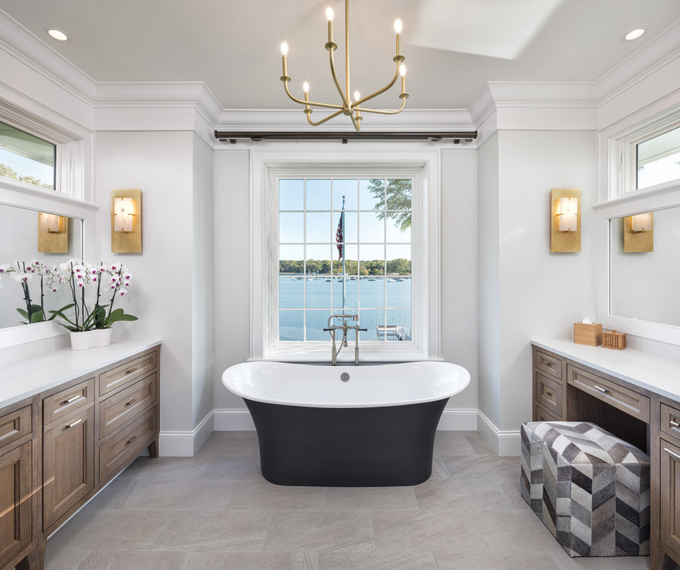 На фото: главная ванная комната в морском стиле с фасадами цвета дерева среднего тона, отдельно стоящей ванной, серыми стенами, серым полом, белой столешницей и зеркалом с подсветкой с