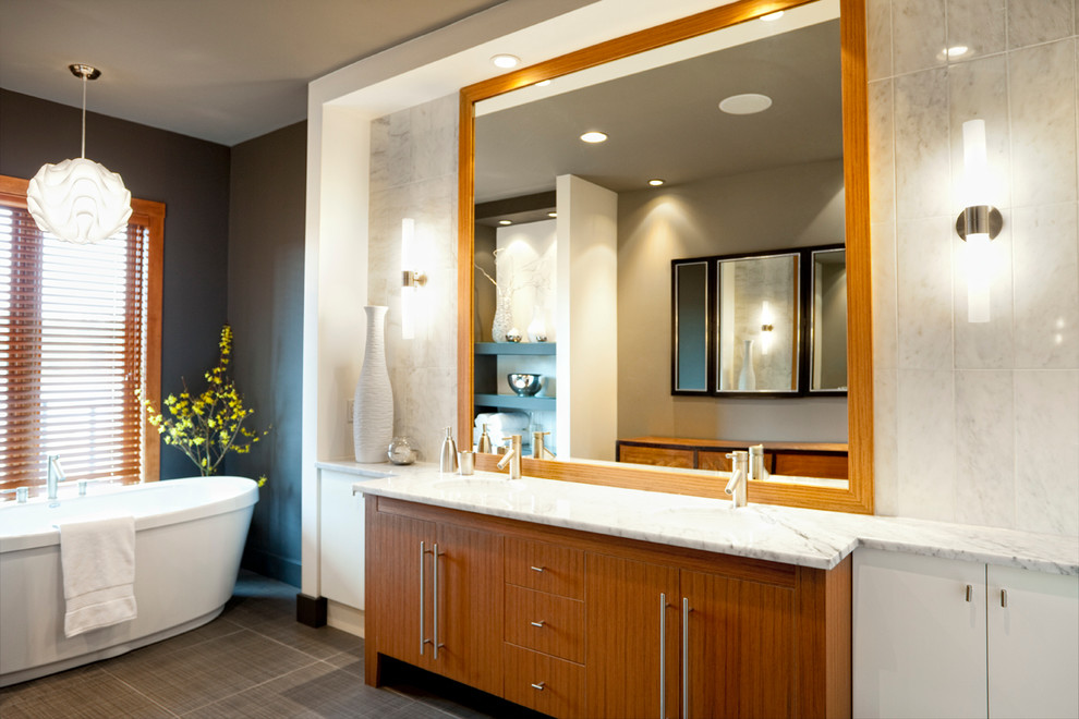 Modern inredning av ett en-suite badrum, med ett fristående badkar, ett undermonterad handfat, släta luckor, vita skåp, marmorbänkskiva, vit kakel, stenkakel, grå väggar och klinkergolv i porslin