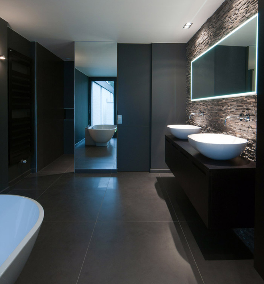 Modern inredning av ett badrum, med skåp i mörkt trä, grå kakel, ett fristående badkar, en vägghängd toalettstol, flerfärgade väggar och klinkergolv i keramik