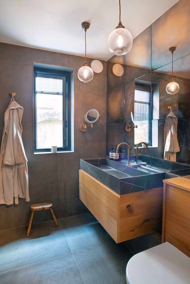Small eclectic ensuite bathroom in London with flat-panel cabinets, medium wood cabinets, a built-in bath, a shower/bath combination, a wall mounted toilet, brown tiles, ceramic tiles, brown walls, ceramic flooring, marble worktops, grey floors, black worktops and a wall-mounted sink.