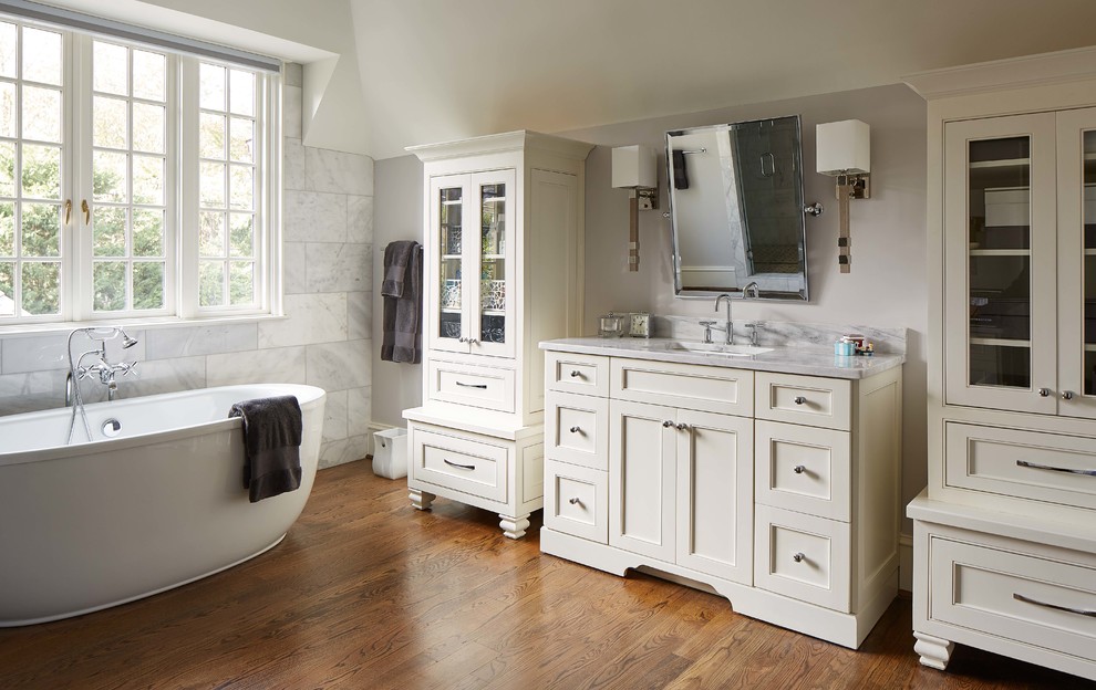 Klassisk inredning av ett en-suite badrum, med ett fristående badkar, vit kakel, grå väggar, mellanmörkt trägolv, ett undermonterad handfat, beige skåp och luckor med infälld panel