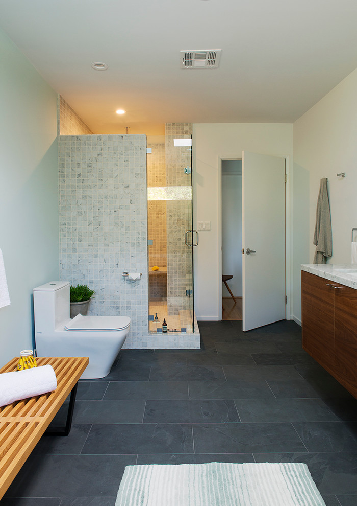 Design ideas for a large modern ensuite bathroom in Los Angeles with a built-in sink, flat-panel cabinets, dark wood cabinets, solid surface worktops, a freestanding bath, a corner shower, a one-piece toilet, black tiles, stone tiles, grey walls, slate flooring, grey floors and a hinged door.
