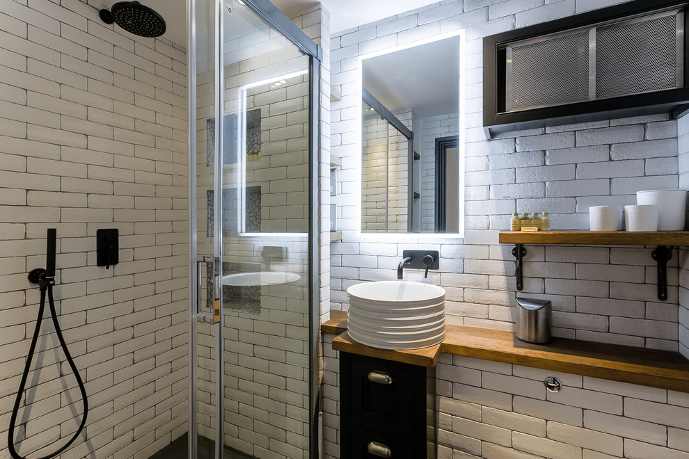 Пример оригинального дизайна: ванная комната в стиле лофт с черными фасадами, душем в нише, белой плиткой, белыми стенами, душевой кабиной, настольной раковиной, столешницей из дерева, черным полом, душем с раздвижными дверями и коричневой столешницей
